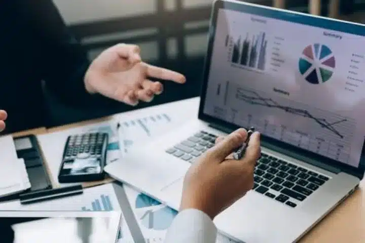 Two professionals discussing financial data displayed on a laptop screen with charts, graphs, and summary reports.
